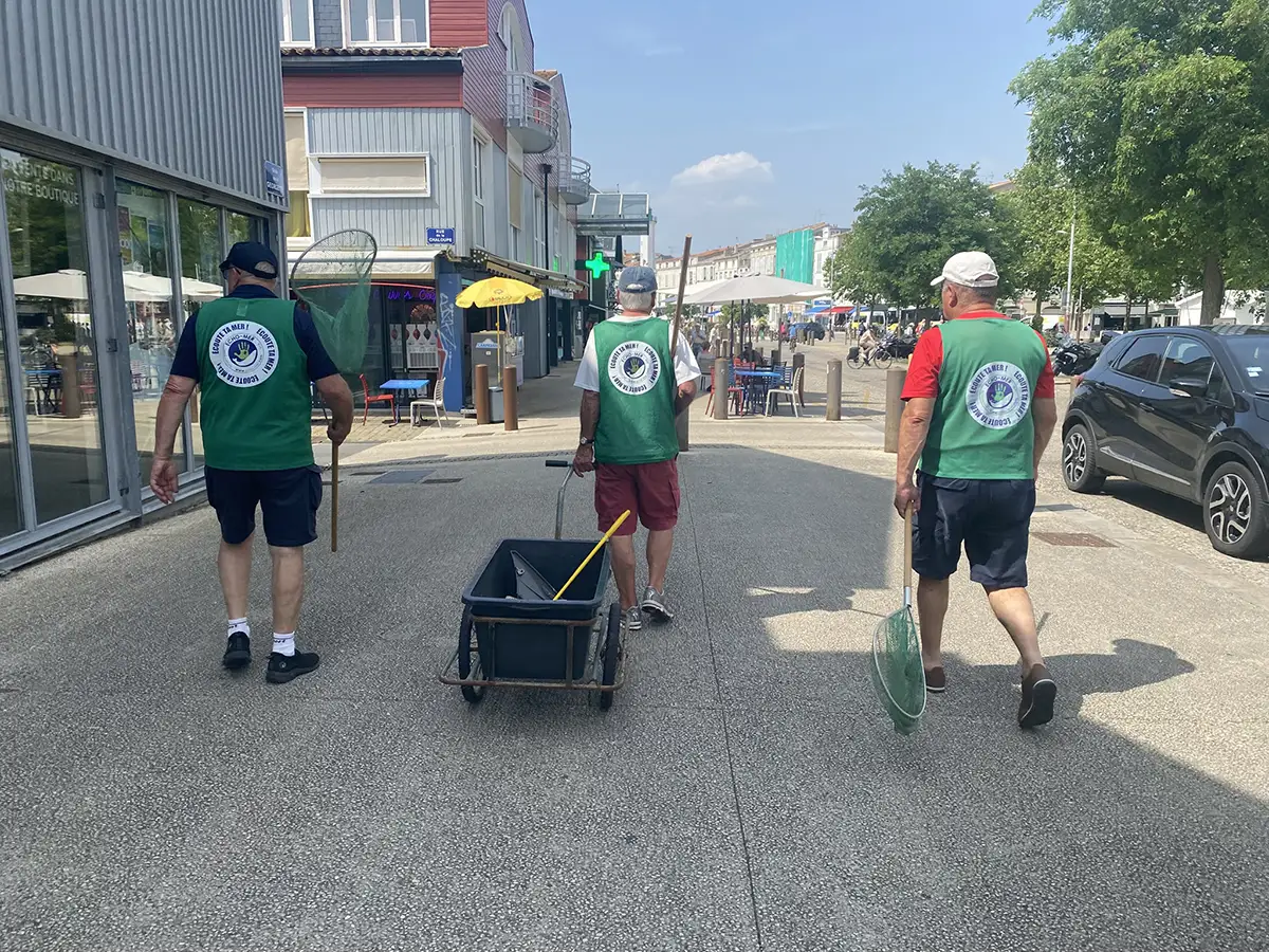 Trois bénévoles en gilet vert, équipés d’épuisettes et d’une brouette, marchent dans une rue piétonne animée près du Bassin des Chalutiers de La Rochelle lors d’une opération de nettoyage.