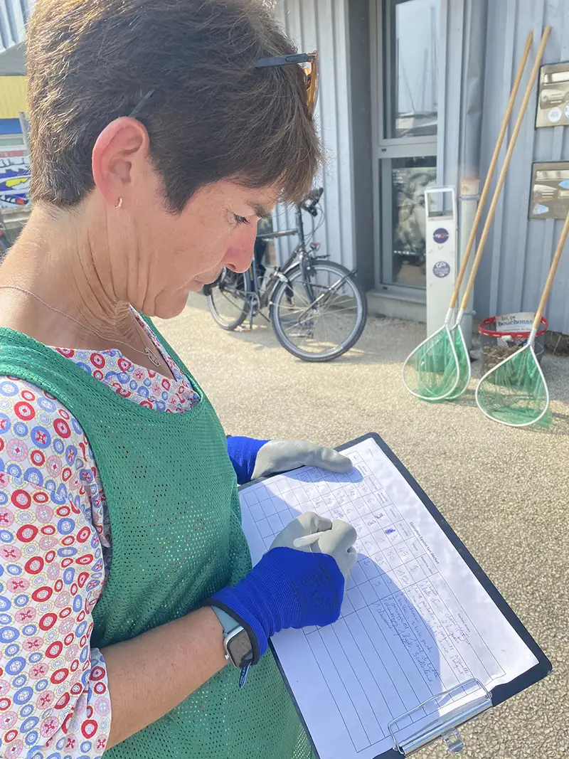 Une bénévole en gilet vert et gants bleus note des informations sur une feuille fixée à un clipboard, lors du suivi de la collecte de déchets.