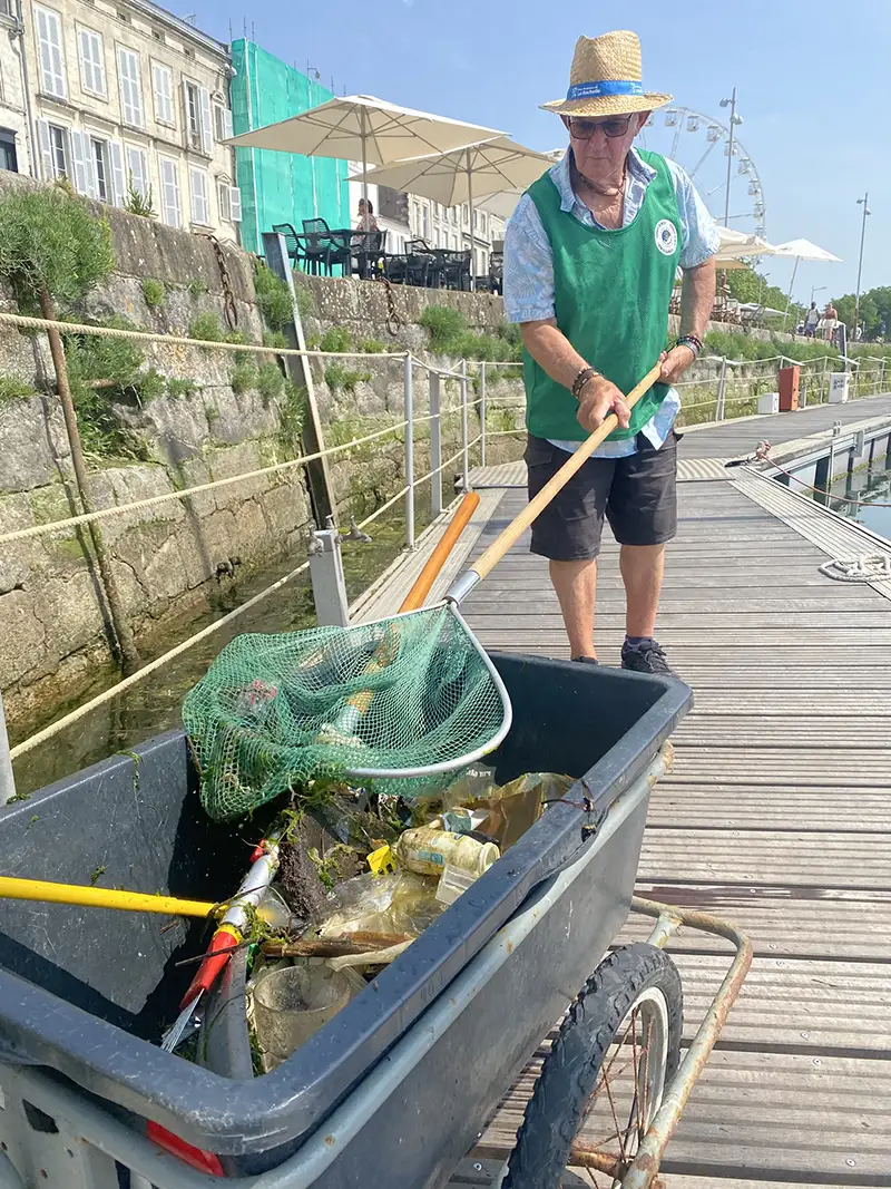 Un bénévole en gilet vert et chapeau de paille ramasse des déchets dans le Vieux-Port de La Rochelle, déposés dans une brouette équipée d’épuisettes et de filets.