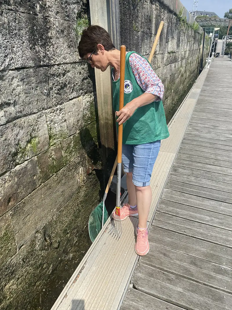 Une bénévole en gilet vert utilise une épuisette pour récupérer des déchets le long du quai dans le port de La Rochelle.